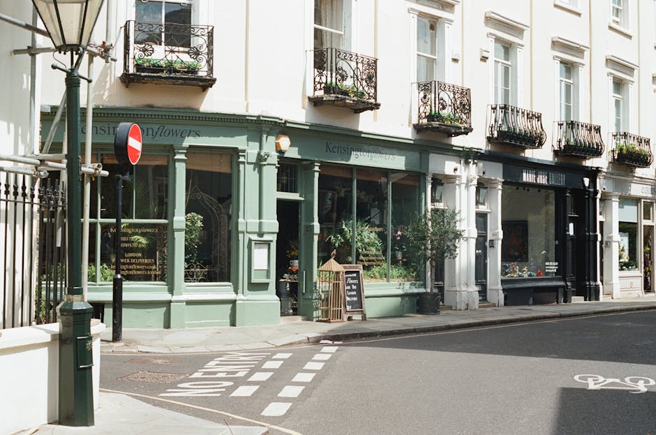 A daytime street scene showing a row of white and light green commercial buildings with large glass windows and decorative black wrought iron balconies on the upper floors, typical of West Kensington architecture. In front of the buildings, a narrow sidewalk features a small chalkboard sign advertising an event or promotion, and a potted plant near the entrance of a flower shop. The street has a designated bike lane marked with white paint and a bicycle symbol, and a one-way traffic sign is mounted on a lamppost at the corner. The lighting is natural, with sunlight casting soft shadows on the pavement and building facades, creating a calm and inviting atmosphere. This environment offers a suitable setting for home relocation or furniture transport services, as showcased by West Kensington Movers' local removals operations, focusing on moving logistics and packing processes within the West Kensington area.