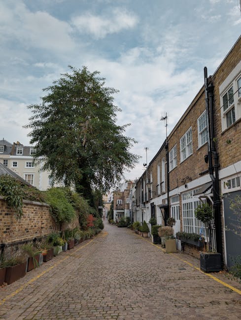 A narrow cobbled street in a residential area, with a gentle curve and lined with multi-story terraced houses featuring white and brick facades, some with small balconies and window boxes filled with